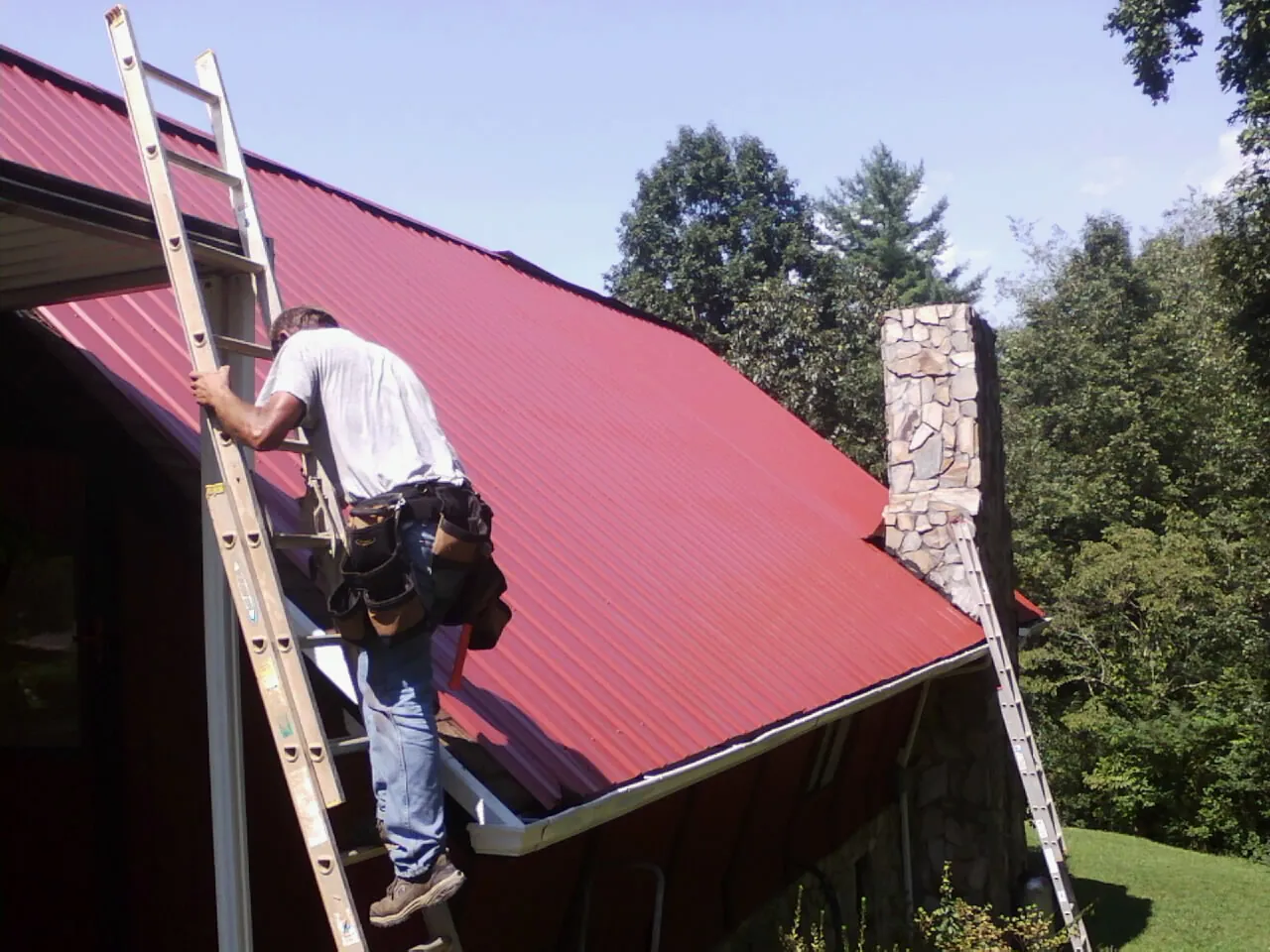 Roofer on a ladder beside a red metal roof and stone chimney — Soffit & Fascia Repair by Roofing Brothers in New Richmond